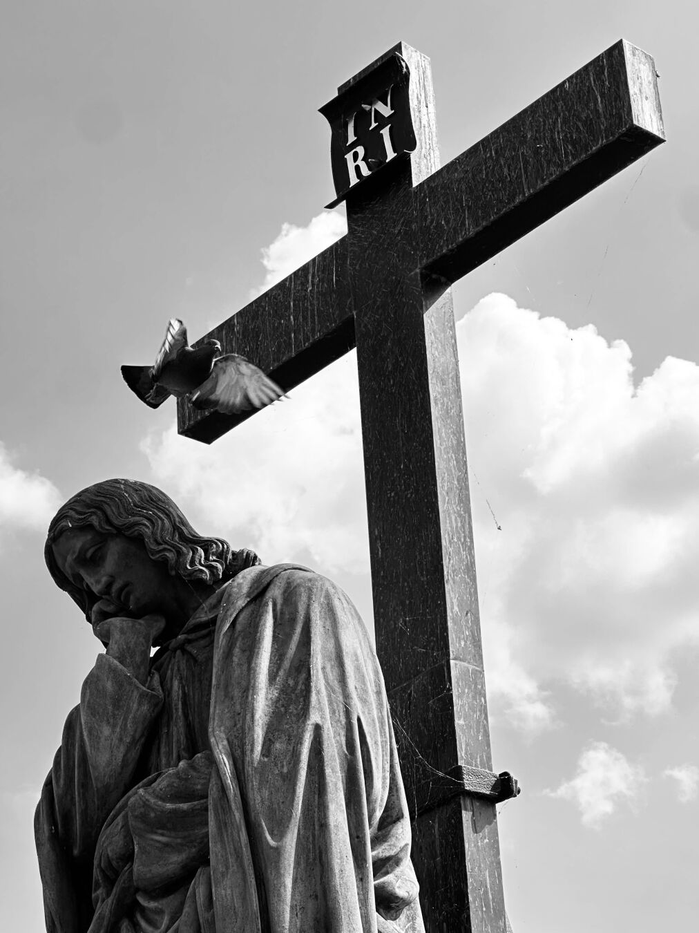 Sotto a una croce con l’iscrizione INRI una statua è in una posizione da facepalm mentre un piccione spicca il volo dalla sua testa